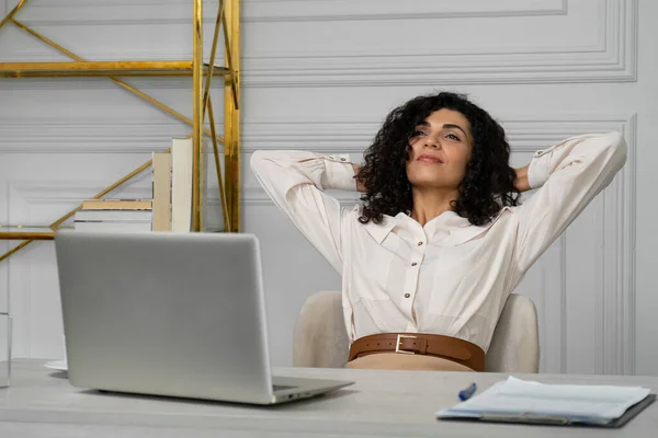 办公室放松的概念。一位印度妇女，一头卷曲的黑发，在笔记本电脑上工作了很长时间，双手放在后脑勺上，坐在一个符合人体工学的办公椅上 — 图库照片