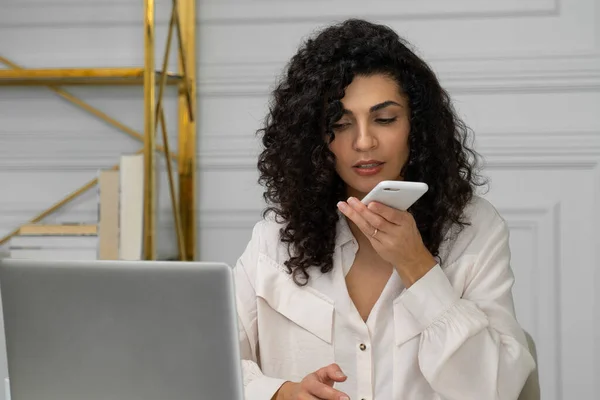 Una joven india enfocada con el pelo negro rizado está grabando un mensaje de voz en su teléfono inteligente, utilizando un asistente de voz digital virtual en un gadget moderno. — Foto de Stock