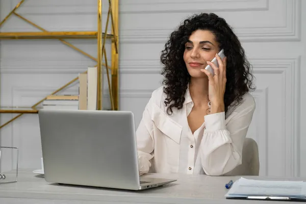 Mujer india joven con el pelo rizado habla en un teléfono móvil mientras trabaja con un ordenador portátil en un escritorio — Foto de Stock