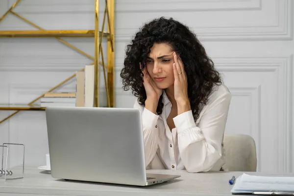Mujer de pelo rizado indio joven y cansado, que sufre de migrañas, sobrecargado de trabajo en un ordenador portátil. Una mujer exhausta se siente mal, toca sus sienes con las manos — Foto de Stock