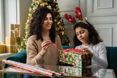 Genç kıvırcık saçlı bir anne küçük kızına bir Noel hediyesi verir. Küçük kız bir Noel ağacının yanında oturan hediyeyi açar.