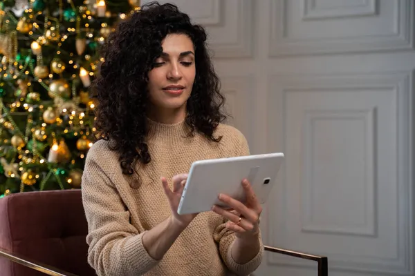 Jonge gekrulde Spaanse vrouw die planeten gebruikt zit thuis op de bank bij de kerstboom.. — Stockfoto