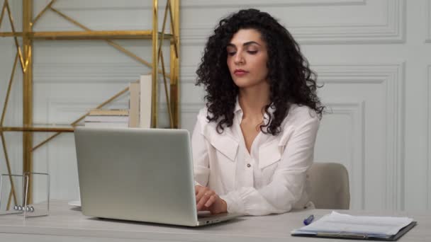 O conceito de relaxamento de escritório. Uma mulher indiana com cabelo preto encaracolado se estende com as mãos atrás da cabeça em uma cadeira de escritório ergonômica depois de trabalhar longas horas em seu laptop — Vídeo de Stock