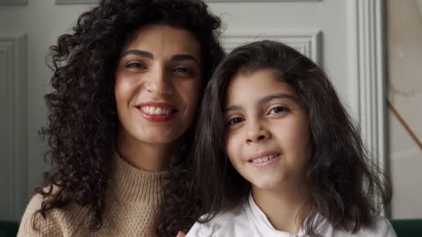 Young, curly-haired mother and her young daughter sit on the couch looking at the camera and smiling — Stock Video