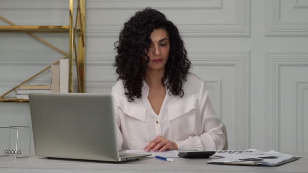 Indian creative curly black-haired woman covers her face with her hand and gets upset from working in front of a laptop on a desk in the office, office lifestyle concept — Stock Video