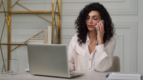 Mulher indiana jovem com cabelo encaracolado fala em um telefone celular enquanto trabalhava com um laptop em uma mesa — Vídeo de Stock