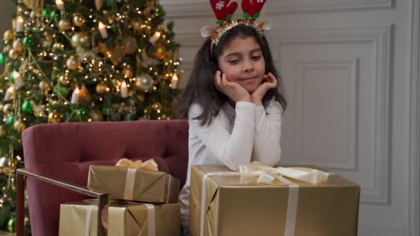 Uma menina pensativa senta-se em uma cadeira com presentes e olha para o fundo de uma árvore de Natal — Vídeo de Stock