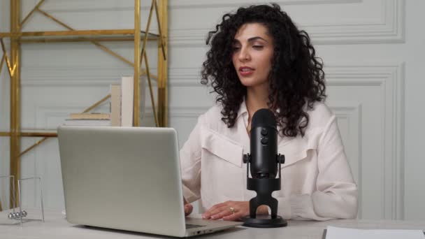 Lockige brünette Frau mit Mikrofon und Laptop, spricht und nimmt im Studio einen Podcast auf — Stockvideo