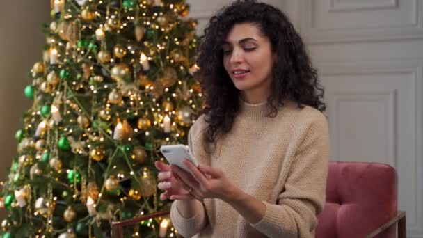 Χαμογελώντας νεαρή γυναίκα χρησιμοποιώντας ένα smartphone, διαβάζοντας τους χριστουγεννιάτικους χαιρετισμούς σε ένα κοινωνικό δίκτυο, κάθεται σε έναν καναπέ κοντά σε ένα διακοσμημένο χριστουγεννιάτικο δέντρο. — Αρχείο Βίντεο