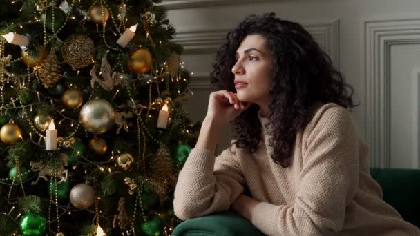 Счастливая, мечтательная, кудрявая темноволосая женщина сидит рядом с елкой, глядя вдаль, загадывая желание. Улыбающаяся молодая женщина наслаждается Рождеством дома, сидя на диване — стоковое видео