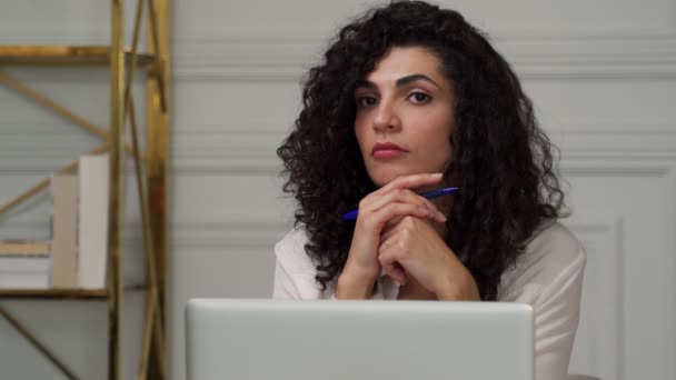 Distratta dal lavoro, una giovane donna indiana con i capelli neri ricci si siede su una scrivania con un computer portatile, pensando. Una donna premurosa guarda fuori dalla finestra con una penna in mano — Video Stock