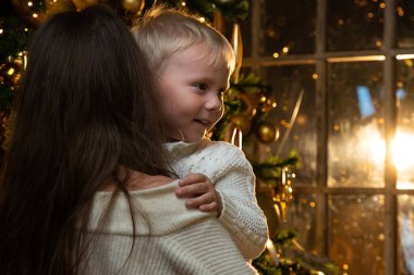Sevgi dolu anne, küçük oğlunu kollarında tutuyor, sarılıyor, Noel ağacının yanında duruyor. Güzel bir kadın bebeğine sarılır. Mutlu yıllar tatili kavramı