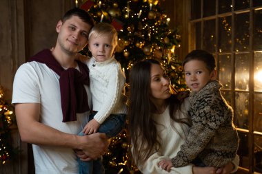 Noel ağacının yanında küçük çocukları olan gülümseyen genç bir ailenin portresi kameraya el sallıyor. Küçük çocuklu mutlu ebeveynler yeni yılı neşeyle kutluyorlar.