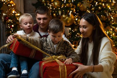 İki küçük oğlu olan mutlu genç bir aile Noel ağacının arka planında Noel hediyelerini açıyor.