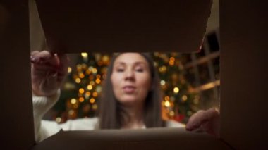 Neşeli, güzel genç bir kadın Noel ağacının arka planında bir Noel hediyesi açıyor. Kutunun içinden görüntüle.