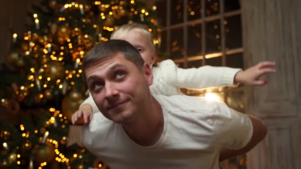 Fiatal apa ad piggyback lovagolni nevetni aranyos kis óvodai gyermek fia, élvezi játszani szórakoztató időt együtt közelében díszített karácsonyfa — Stock videók