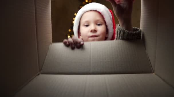 Niño feliz en un sombrero de Santa Claus mira en una caja de regalo. Un niño desempaca una caja con un regalo de Navidad. Vista desde la caja — Vídeo de stock