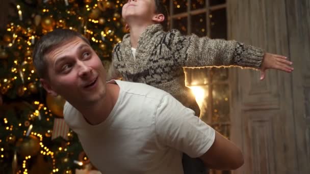Slow-motion young father giving piggyback ride to laughing cute little preschool child son, enjoying playing spending fun time together near decorated Christmas tree — Stock Video