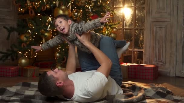 Le père se trouve sur le sol chaud en bois dans le salon, décoré d'un sapin de Noël lumineux, ramasse son petit fils, profitant du concept de bonnes vacances — Video