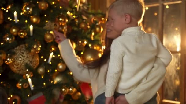 Junge Mutter in weißer Bluse schmückt mit ihrem Sohn einen Weihnachtsbaum — Stockvideo