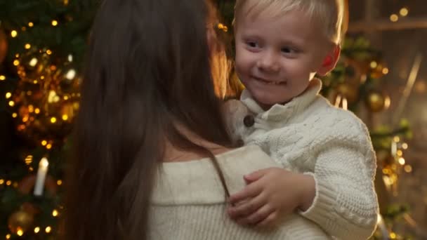 Szerető anya a karjaiban tartja a kisfiát, ölelkezik, a karácsonyfa mellett áll. Egy csinos nő megöleli a gyerekét. A boldog újévi ünnepek koncepciója — Stock videók