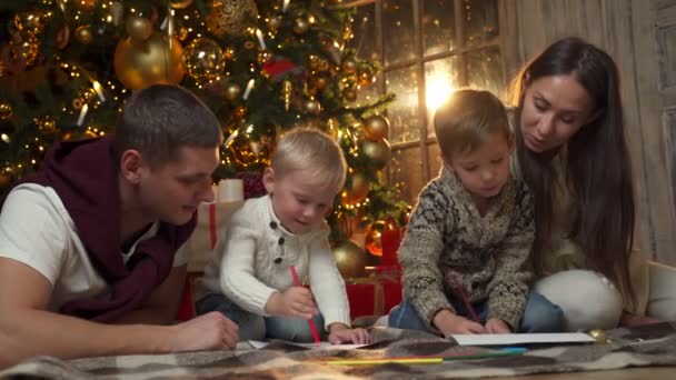 De jeunes parents avec deux jeunes fils dessinent assis sur le sol. La famille célèbre, passe du temps ensemble la veille de Noël ou le réveillon du Nouvel An — Video