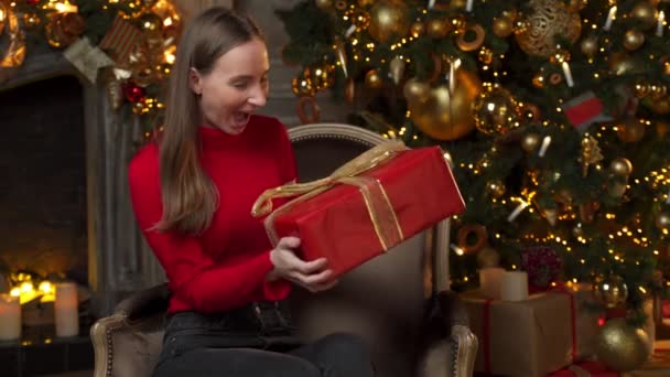 Gelukkig vrouw in een rode trui opent een magische kerstcadeau thuis op de achtergrond van een kerstboom — Stockvideo