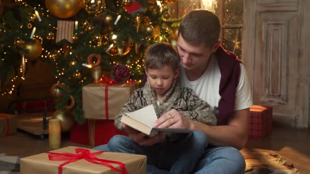 Babalık kavramı, mutlu bir baba ve küçük oğlunun Noel ağacının yanında, evin zemininde oturup kitap okumasıdır. — Stok video