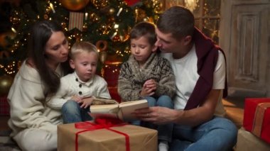 Mutlu genç bir aile ve iki oğlu Noel ağacında yerde oturmuş kısa hikayeler okuyorlar..