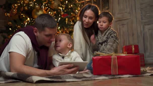 행복 한 어린 부모와 어린 자녀가 크리스마스 트리 근처의 따뜻 한 바닥에 앉아 알약을 사용하여 친척들 과 비디오 통화를 하고 있습니다 — 비디오