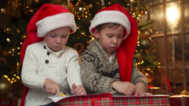 Dzieci piszą listy do Świętego Mikołaja siedzącego przy choince. Dwóch małych braci na podłodze na tle choinki — Wideo stockowe