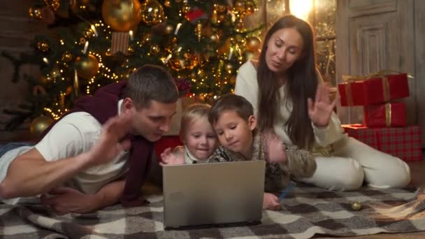 Mladá rodina a dva mladí synové pořádají na Štědrý večer videokonferenci na Skypu, mávají rukama a blahopřejí příbuzným k používání notebooku na pozadí vánočního stromečku — Stock video