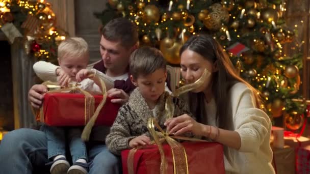Boldog fiatal család két kisfiúval együtt nyitott karácsonyi ajándékok a háttérben egy karácsonyfa — Stock videók
