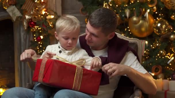 Alegre padre abraza a su pequeño hijo encantador, la familia celebra la Navidad, abre regalos, se sienta en una silla cerca del árbol de Navidad — Vídeos de Stock