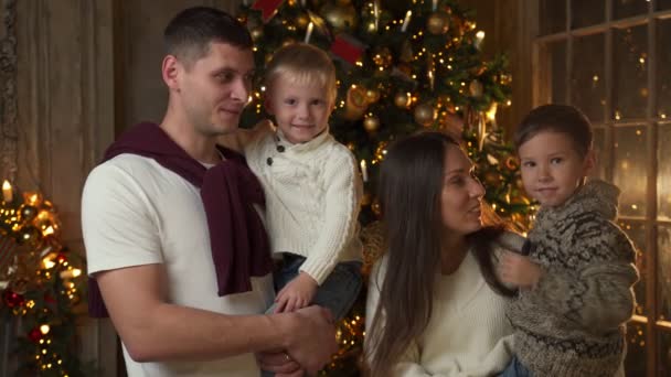 Porträtt av en leende ung familj med små barn nära julgranen viftar en hälsning till kameran. Glada föräldrar med små barn firar glatt nyår semester — Stockvideo