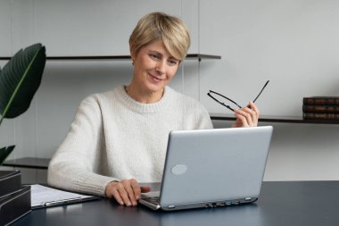 Ofiste dizüstü bilgisayarda çalışan olgun bir iş kadınının portresi, gözlüğünü çıkarıyor ve dizüstü bilgisayarına bakarken düşünüyor.