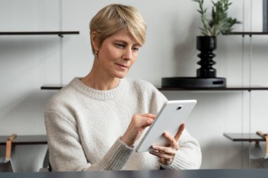 Orta yaşlı, gülümseyen beyaz bir kadının portresi. Bir koltukta oturuyor, modern bir tablet kullanıyor, internetten iletişim kuruyor.