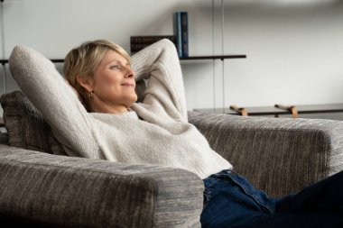 Olgun kadın evinde bir sandalyede dinleniyor. Orta yaşlı bir kadın pencerenin yanındaki modern bir sandalyeye oturmuş oturma odasında dinleniyor.