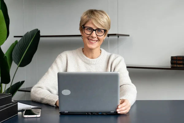 Уверенная женщина средних лет, работающая за ноутбуком в современном офисе, смотрит в камеру — стоковое фото