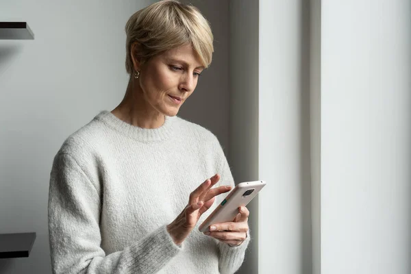 Encantadora mujer de mediana edad de pie junto a la ventana y escribiendo mensajes de texto en su teléfono, utilizando un teléfono inteligente moderno en Internet — Foto de Stock