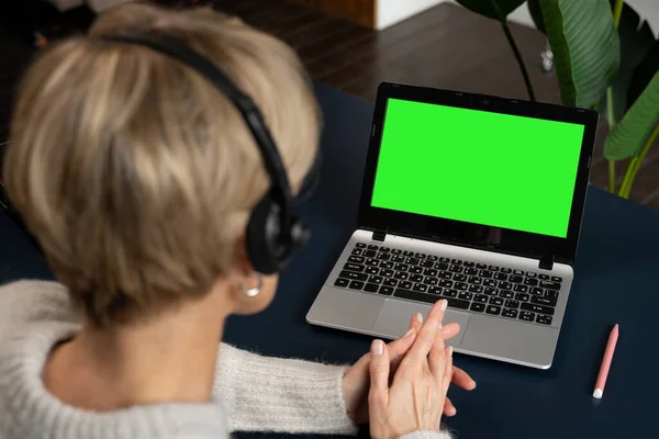 Widok przez ramię kobiety w średnim wieku w słuchawkach, pracującej lub studiującej na laptopie z zielonym ekranem. — Zdjęcie stockowe