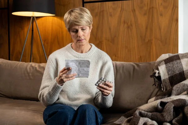 Mujer de mediana edad sostiene un paquete de pastillas y lee instrucciones médicas sentadas en el sofá — Foto de Stock