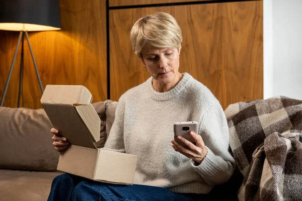 Mujer de mediana edad sentada en el sofá abre la caja, comprueba el paquete con un teléfono inteligente — Foto de Stock