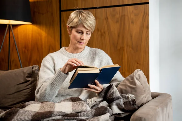 Mooi middelbaar blond is het lezen van een boek thuis zitten op de bank — Stockfoto
