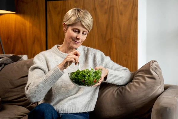 Женщина средних лет сидит и отдыхает дома на диване, ест зеленый салат. Женщина, питающаяся здоровой пищей. — стоковое фото