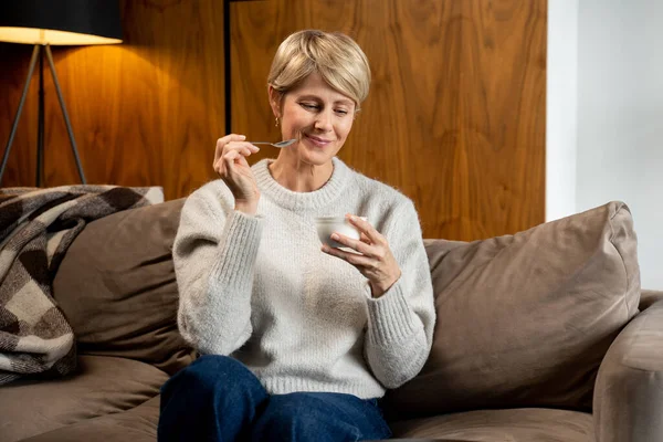 Retrato de una mujer de mediana edad que come yogur sentado en el sofá en casa — Foto de Stock