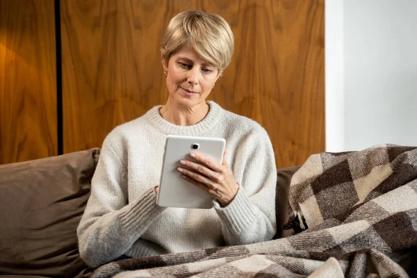 Retrato de una mujer de mediana edad sentada en un sofá acogedor y usando una tableta en casa. Una mujer de compras en una popular tienda online — Foto de Stock