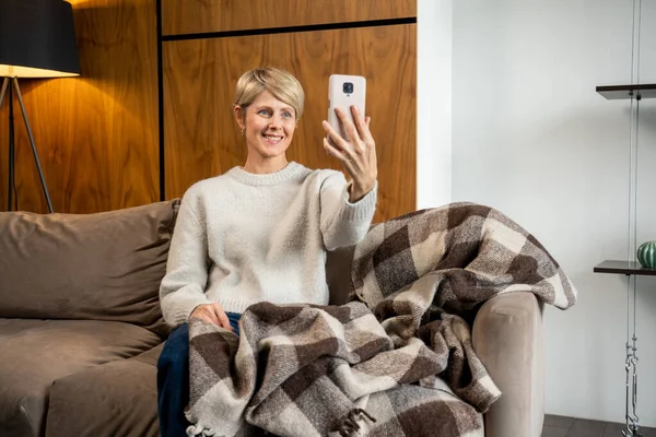 Una mujer de mediana edad está sentada en el sofá de la sala de estar, hablando por video en un gadget de teléfono celular moderno, una mujer mayor saludando, hablando en Internet usando un teléfono inteligente — Foto de Stock
