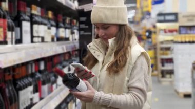 Genç bir kadın akıllı telefonunu kullanarak bir şişe şarabın şifresini tarıyor. Arka planda bir süpermarket var..