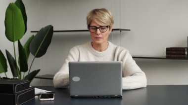 Modern bir ofiste dizüstü bilgisayarda çalışan kendinden emin orta yaşlı bir kadın kameraya bakar.